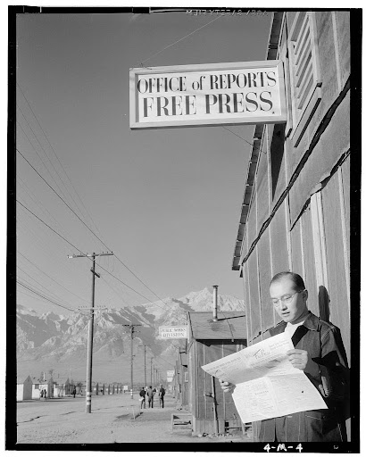 A black and white photo taken by Ansel Adams in a Japanese internment camp during World War II titled 'Roy Takeno reading paper in front of office'